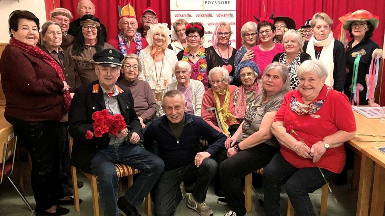 A group of elderly people dressed in various costumes and hats are posing for a photo. They are seated and standing, with a banner in the background.
