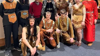 Eine Gruppe von Menschen in indianischer Kleidung posiert für ein Foto, mit einer Frau in Rot und einem Mann mit Hut in der Mitte.