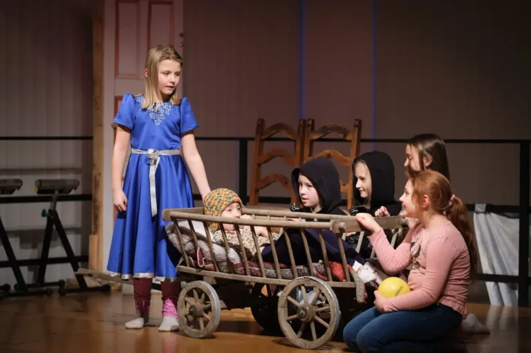 A girl in a blue dress stands beside a wooden cart with a baby inside, while several children sit around it. One child holds a yellow ball. They are on a stage.