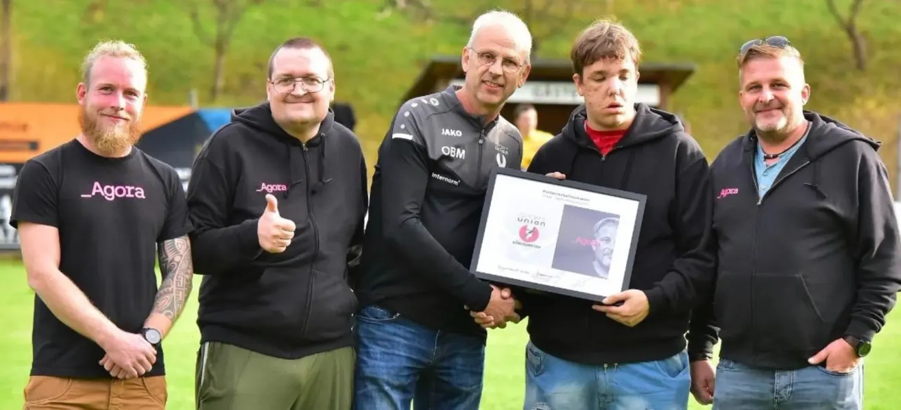 Three men stand together on a field. The man in the middle holds a framed certificate. The man on the left gives a thumbs up. The man on the right looks at the man in the middle.