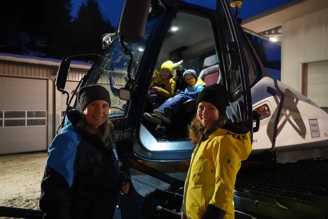 Two women and two kids are standing in front of a snow plow vehicle. The two kids are sitting inside the vehicle. The women are smiling. The kids are wearing hats.