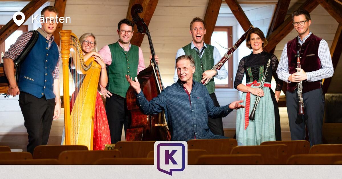 Fünf Musiker, jeder mit einem anderen Instrument, stehen lächelnd zusammen. Die Instrumente sind Harfe, Bass, Fagott, Klarinette und Cello. Sie befinden sich in einem Raum mit Holzbalken und einem Fenster.