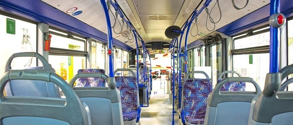 Ein leerer Bus mit blauen und grauen Sitzen. Es gibt blaue Haltestangen, einen Monitor und einen Notfallgriff an der Decke.