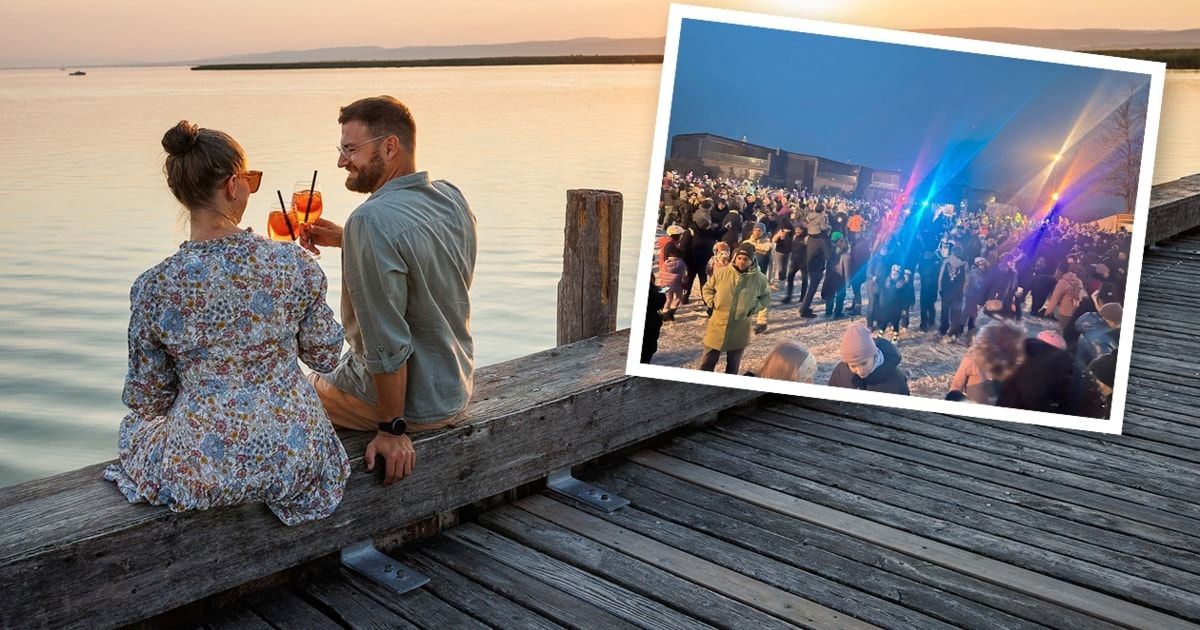 Ein Mann und eine Frau sitzen auf einem Holzsteg am Meer und halten Getränke, während eine Menschenmenge bei einer Veranstaltung am Strand bei Nacht versammelt ist.