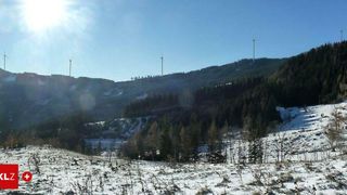 Eine Landschaft mit klarem blauem Himmel, verschneiten Bergen und Windturbinen auf den Graten. Bäume bedecken die unteren Hänge, und der Vordergrund ist mit Schnee bedeckt.