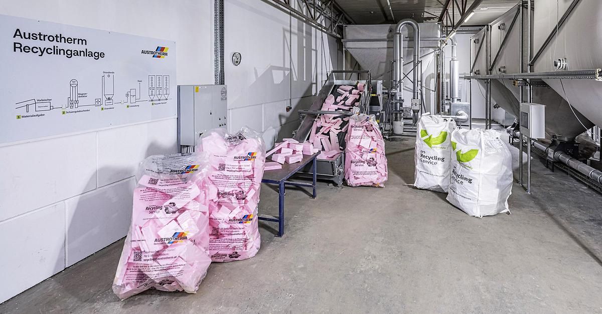 Ein Industriebereich mit mehreren rosafarbenen Säcken, die mit 'Recycling-Service' beschriftet sind, und einem Förderband mit rosa Objekten. Eine weiße Wand hat ein Diagramm und ein Schild, das 'AUSTROTERM' liest. Es gibt einen Tisch mit Säcken und einen großen weißen Sack auf dem Boden.