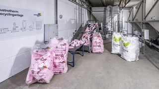 Ein Industriebereich mit mehreren rosafarbenen Säcken, die mit 'Recycling-Service' beschriftet sind, und einem Förderband mit rosa Objekten. Eine weiße Wand hat ein Diagramm und ein Schild, das 'AUSTROTERM' liest. Es gibt einen Tisch mit Säcken und einen großen weißen Sack auf dem Boden.
