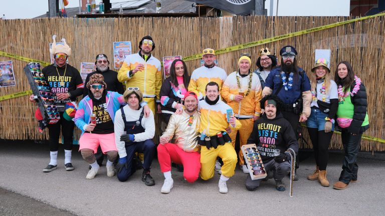 Eine Gruppe von Menschen, die in übereinstimmenden gelben und weißen Kostümen mit Sonnenbrillen und Mützen gekleidet sind, posiert für ein Foto. Sie stehen vor einem Bambuszaun.