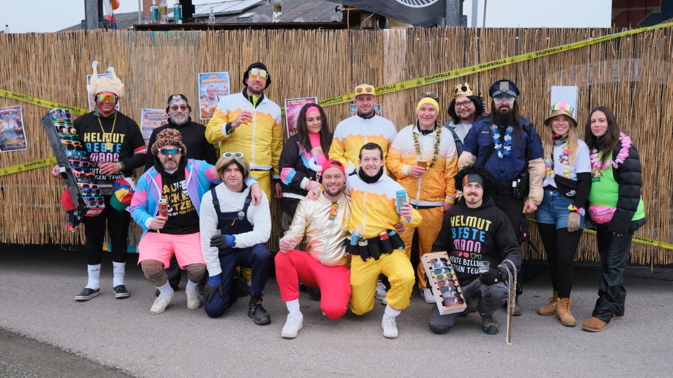 Eine Gruppe von Menschen, die in übereinstimmenden gelben und weißen Kostümen mit Sonnenbrillen und Mützen gekleidet sind, posiert für ein Foto. Sie stehen vor einem Bambuszaun.