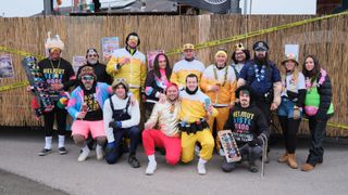 Eine Gruppe von Menschen, die in übereinstimmenden gelben und weißen Kostümen mit Sonnenbrillen und Mützen gekleidet sind, posiert für ein Foto. Sie stehen vor einem Bambuszaun.