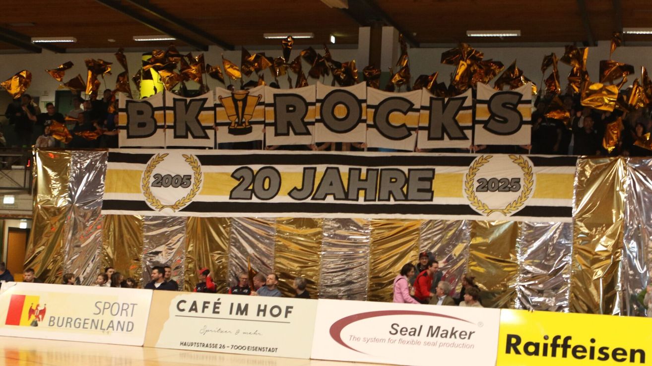 Ein Feierbanner mit der Aufschrift 'BK Rocks 20 Jahre' von 2005 bis 2025. Zuschauer sitzen auf Tribünen darunter, einige tragen Mützen und Jacken. Banner und goldene Girlanden schmücken den Hintergrund.