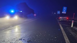 Eine dunkle Straße bei Nacht mit zerbrochenem Glas auf dem Boden und einem Fahrzeug in der Ferne mit eingeschalteten Scheinwerfern.