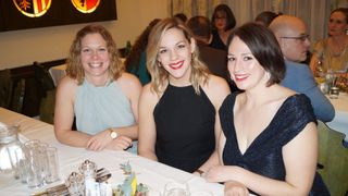 Drei Frauen lächeln und posieren für ein Foto. Die Frau auf der linken Seite trägt ein blaues ärmelloses Top und eine Uhr. Die Frau in der Mitte trägt ein schwarzes Kleid mit rotem Lippenstift. Die Frau auf der rechten Seite trägt ein schwarzes Kleid mit einem V-Ausschnitt.