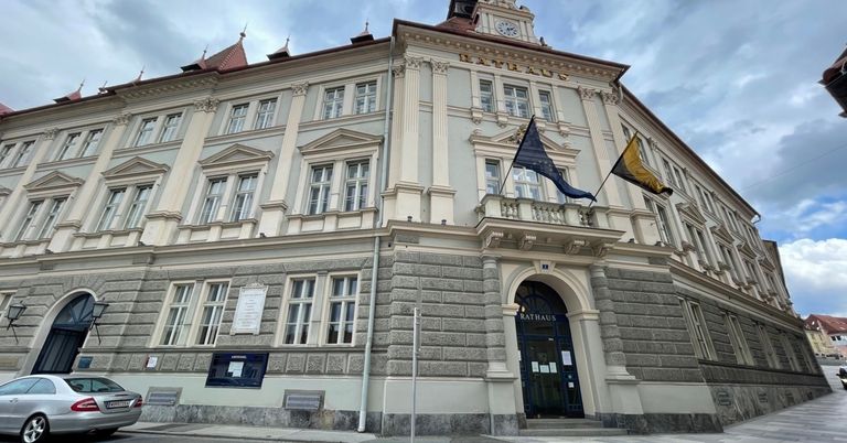 Ein großes, klassizistisches Gebäude mit mehreren Fenstern und einem Uhrturm. Zwei Flaggen, eine blau und eine gelb, sind auf dem vorderen Balkon angebracht.
