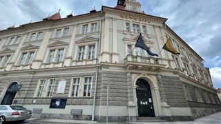 Ein großes, klassizistisches Gebäude mit mehreren Fenstern und einem Uhrturm. Zwei Flaggen, eine blau und eine gelb, sind auf dem vorderen Balkon angebracht.
