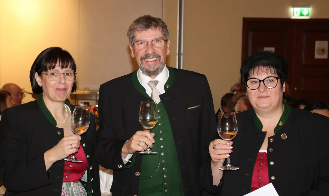 Ein Mann und zwei Frauen stehen in einem Raum und halten Weingläser. Sie lächeln und der Mann trägt eine grüne Weste mit Knöpfen. Die Frauen tragen schwarze Mäntel.