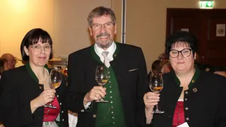 Ein Mann und zwei Frauen stehen in einem Raum und halten Weingläser. Sie lächeln und der Mann trägt eine grüne Weste mit Knöpfen. Die Frauen tragen schwarze Mäntel.