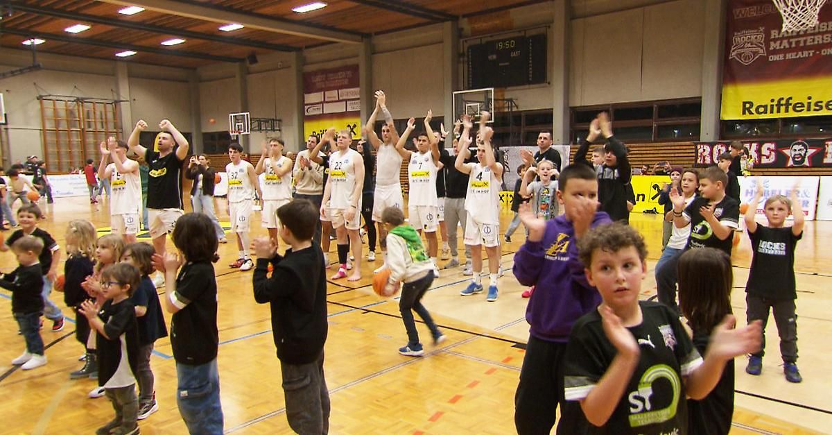 A basketball game with players in white jerseys and spectators in the stands. Some players have raised hands while others hold a basketball. There is a digital scoreboard above the court.