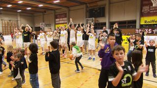 Ein Basketballspiel mit Spielern in weißen Trikots und Zuschauern in den Rängen. Einige Spieler haben erhobene Hände, während andere einen Basketball halten. Über dem Spielfeld befindet sich ein digitales Anzeigebrett.