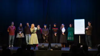 Eine Gruppe von Menschen, darunter eine Frau mit einem Mikrofon, steht auf einer Bühne vor einem blauen Vorhang. Rechts befindet sich eine Tafel mit Schrift. Sie stehen möglicherweise auf der Bühne.