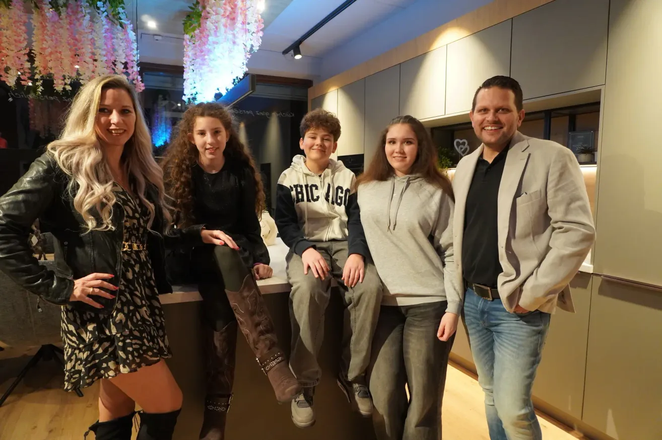 Eine Familie mit fünf Personen, darunter zwei Erwachsene und drei Kinder, posiert für ein Foto in einem modernen Interieur. Die Erwachsenen stehen auf beiden Seiten, und die Kinder sitzen auf einer Theke. Das Interieur hat weiße Wände und einen Holzboden.