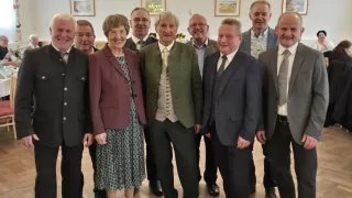 Eine Gruppe von Menschen, möglicherweise eine Familie, posiert für ein Foto in einem Raum mit weißen Wänden. Sie tragen alle formelle Kleidung. Die Frau in der Mitte lächelt und schaut in die Kamera. Hinter ihnen hängen Bilderrahmen an der Wand.