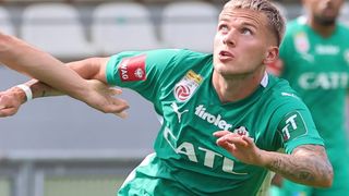 Ein Fußballspieler in einem grünen Trikot mit dem Wort 'tirler' darauf wird von einem anderen Spieler getackelt. Er hat einen ernsten Gesichtsausdruck.