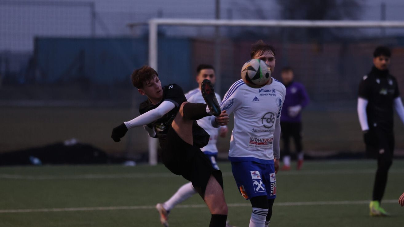 Zwei Fußballspieler sind auf einem Feld in Aktion. Ein Spieler tritt den Ball, während der andere versucht, ihn zu blockieren. Beide tragen Trikots mit Logos. Im Hintergrund ist ein Torpfosten zu sehen.