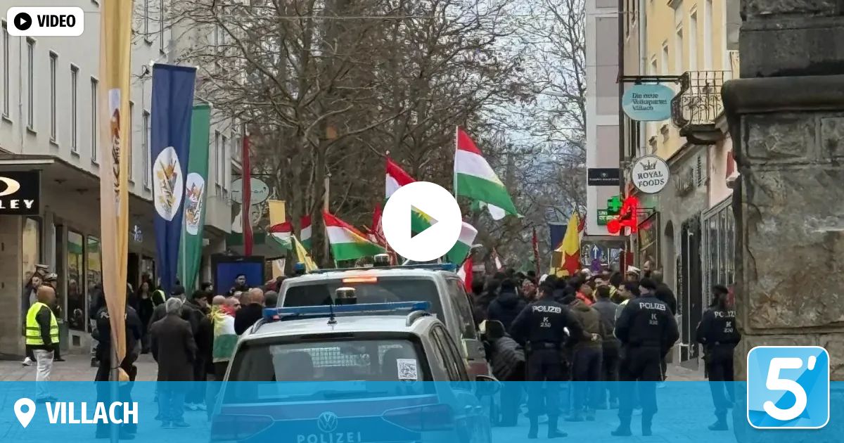 Eine Menschenmenge versammelt sich auf einer Stadtstraße, wo verschiedene Flaggen, einschließlich der ungarischen Flagge, gezeigt werden. Polizeibeamte sind anwesend und Autos sind entlang der Straße geparkt.