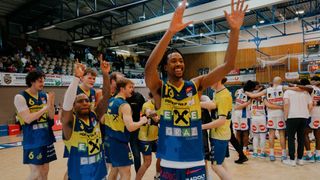 Basketballspieler feiern in einer Turnhalle mit blauen und gelben Uniformen, ein Spieler vorne trägt die Nummer 13 und hebt beide Hände.