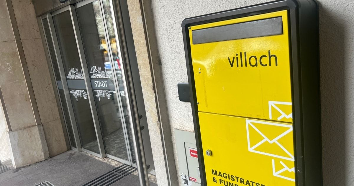 Ein gelber Briefkasten mit dem Wort 'Villach', der an einer weißen Wand neben einer Glastür montiert ist. Die Glastür spiegelt ein Gebäude und ein Auto wider.