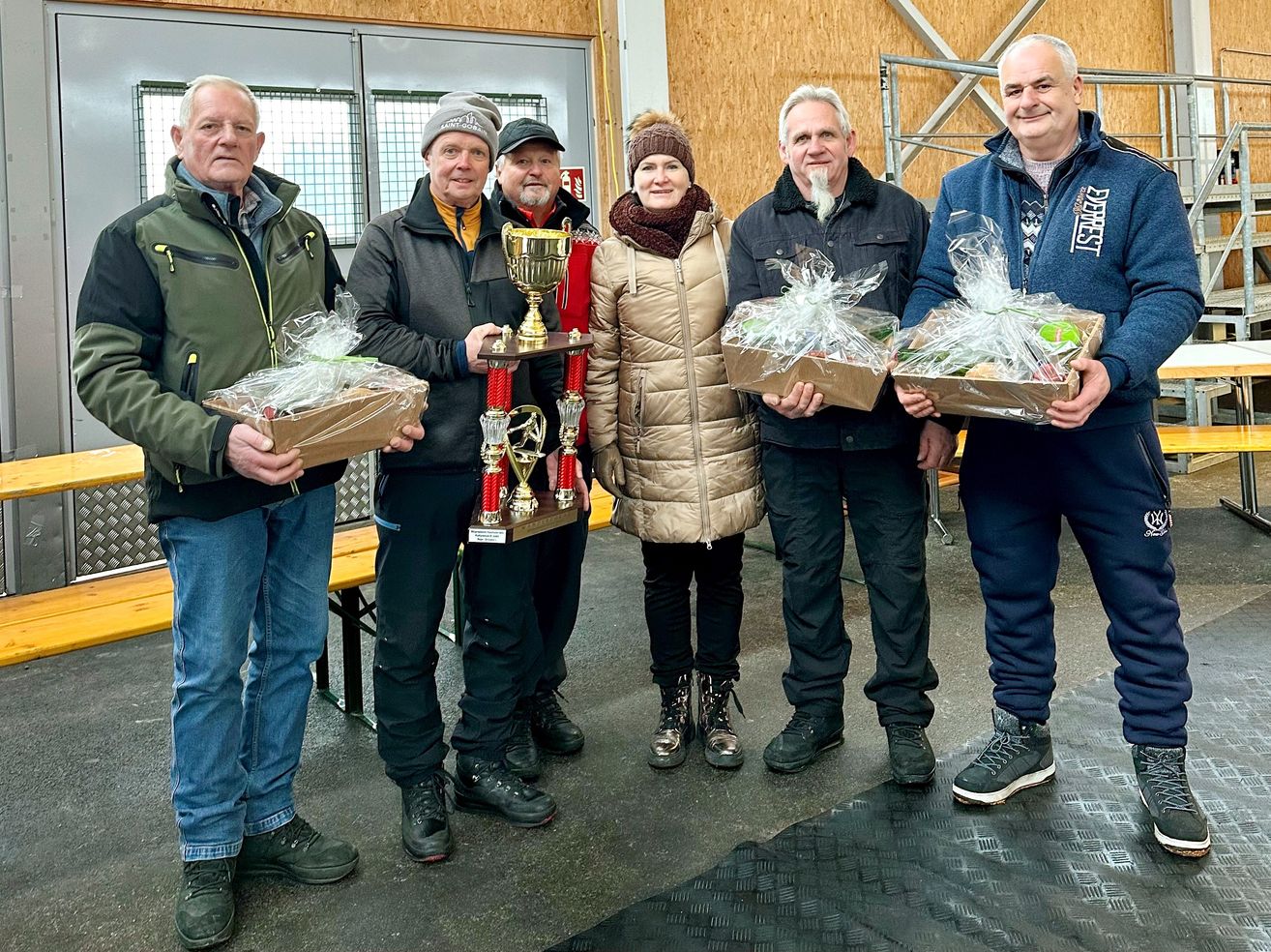 Fünf Menschen stehen in einem Raum zusammen und halten Pokale und Geschenkkörbe. Ein Mann hat einen Pokal, die anderen haben Geschenkkörbe. Die Frau in der Mitte trägt Hut und Schal. Hinter ihnen steht eine Holzbank.