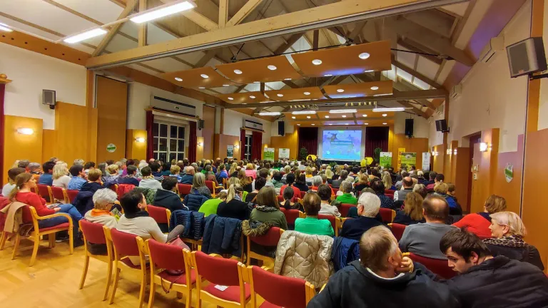 Eine große Gruppe von Menschen ist in einem Konferenzsaal versammelt und hört sich eine Präsentation auf einer Projektionsleinwand an. Der Raum hat Holzbalken, rote Stühle und einen Holzboden.