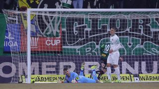 Ein Fußballspieler liegt vor einem Tor am Boden, trägt ein blaues Trikot und Handschuhe. Ein anderer Spieler steht hinter dem Torpfosten und lächelt. Ein dritter Spieler steht in der Nähe des Tores.