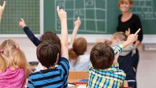 Ein Klassenzimmer mit Kindern, die ihre Hände heben, um eine Frage zu beantworten, eine Lehrerin vorne mit einer grünen Tafel im Hintergrund.