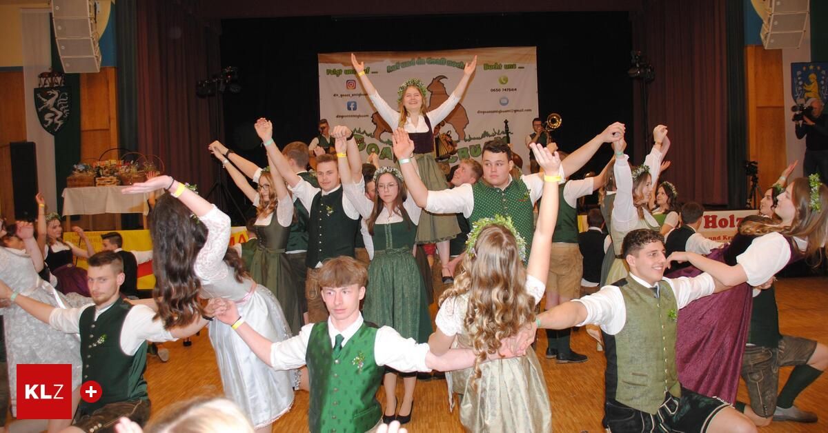 Eine Gruppe von Menschen in traditioneller Kleidung tanzt auf der Bühne, einige halten sich an den Händen, während andere die Arme heben. Ein Banner ist hinter ihnen angebracht.