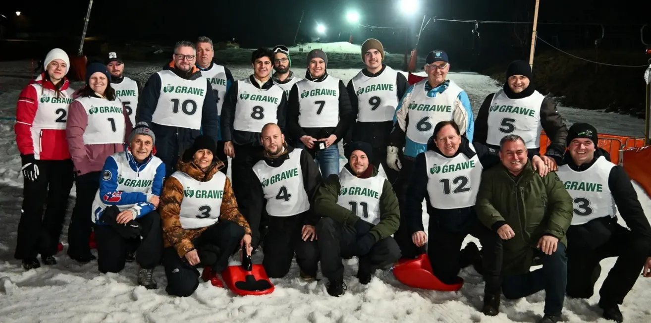 A group of people are wearing numbered vests, smiling, and posing for a photo in the snow. They are holding red sleds. The background is snowy with lights.