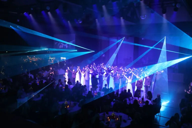 Eine lebhafte Aufführung auf einer Bühne mit blauer Beleuchtung, bei der mehrere Menschen in Gruppen tanzen. Einige sind in weißen Outfits, andere in Militäruniformen. Das Publikum sitzt vor ihnen und genießt die Show.