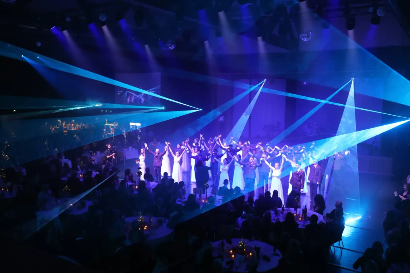 Eine lebhafte Aufführung auf einer Bühne mit blauer Beleuchtung, bei der mehrere Menschen in Gruppen tanzen. Einige sind in weißen Outfits, andere in Militäruniformen. Das Publikum sitzt vor ihnen und genießt die Show.