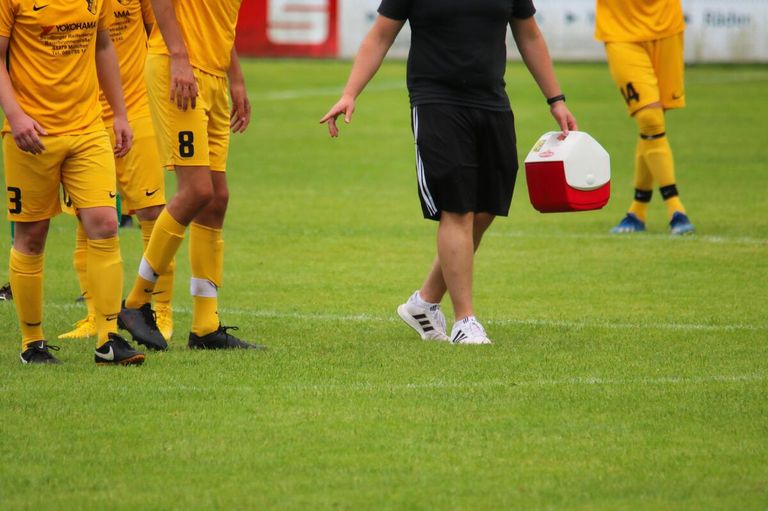 Soccer players in yellow uniforms and a man in black carrying a cooler walk on a green field.