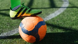 Der grüne und schwarze Schuh eines Fußballspielers liegt auf einem orangefarbenen Fußball mit blauen Mustern auf einem grünen Spielfeld.
