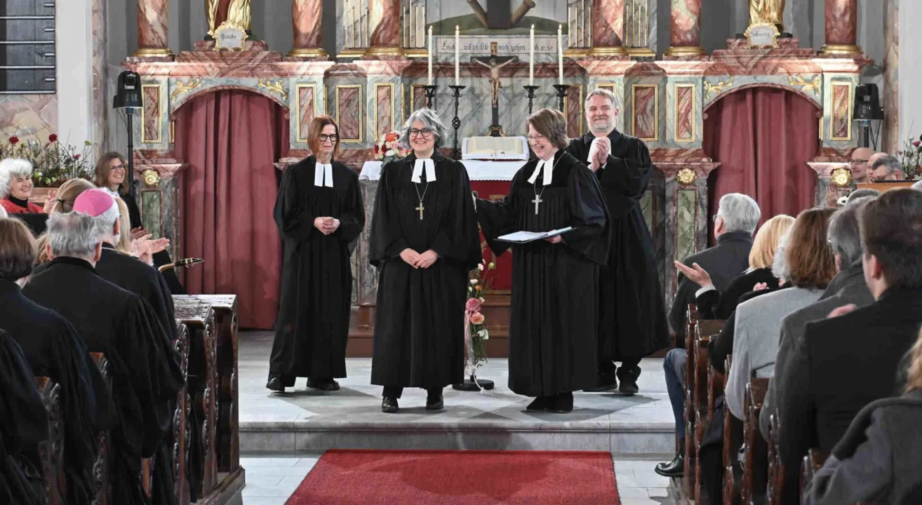 Vier Personen in schwarzen Roben stehen auf einer Kirchenbühne. Sie halten Papiere und tragen Brillen und Halsketten. Die Bühne hat Kerzen, Blumen und einen roten Teppich. Ein Kreuz und eine Statue sind im Hintergrund.