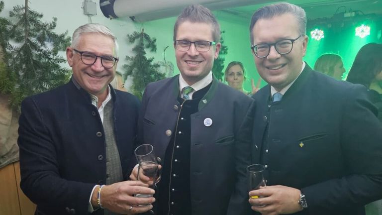 Drei Männer, gekleidet in formeller Kleidung, lächeln für die Kamera. Sie halten Gläser in den Händen, und einer trägt eine grüne Krawatte. Eine Frau steht im Hintergrund, und ein Baum ist links zu sehen.