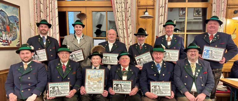 A group of men dressed in traditional attire pose for a photo, holding framed awards. They are in a room with wooden walls, curtains, and lamps.