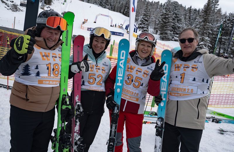 Vier Personen stehen zusammen auf einem verschneiten Hang, lächeln für die Kamera. Sie halten Skier und tragen nummerierte weiße Shirts. Hinter ihnen ist ein verschneiter Hügel mit Bäumen und einer Fahne zu sehen.