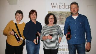 Vier Personen stehen lächelnd vor einer Wand. Zwei halten Weinflaschen, eine hat ein Weinglas und eine weitere hat eine Weinflasche und ein Glas. An der Wand hängt ein Banner.