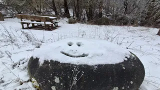 Ein schneebedeckter Felsen mit einem lächelnden Gesicht steht in einem verschneiten Park mit einer Holzbank und einem Tisch, umgeben von Bäumen.