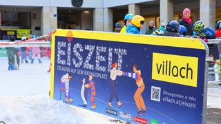 Ein buntes Schild, das Eislaufen auf dem Rathausplatz bewirbt, mit einer Illustration von Menschen beim Eislaufen und einem QR-Code. Es wird in einem verschneiten Bereich mit Menschen in Helmen angezeigt.