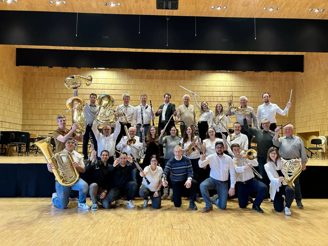 Eine Band in formeller Kleidung, bestehend aus Trompeten, Posaunen und Saxophonen, posiert für ein Foto in einem Musiksaal.