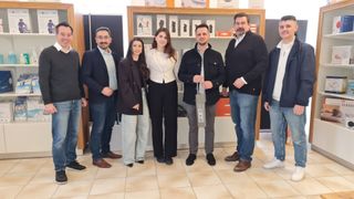 Eine Gruppe von Menschen, darunter zwei Frauen und vier Männer, steht in einem Raum zusammen. Ein Mann hält einen Papierbeutel. Dahinter befindet sich ein Regal mit Produkten.